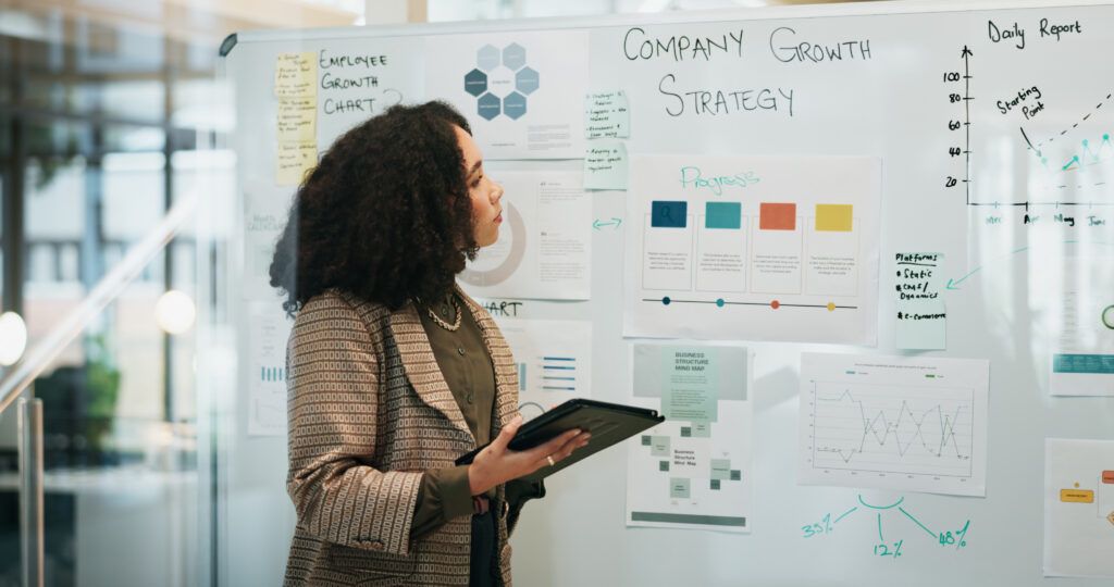 Woman in a blazer studies a whiteboard covered with charts, graphs, and sticky notes from a strategic planning session (side profile).
