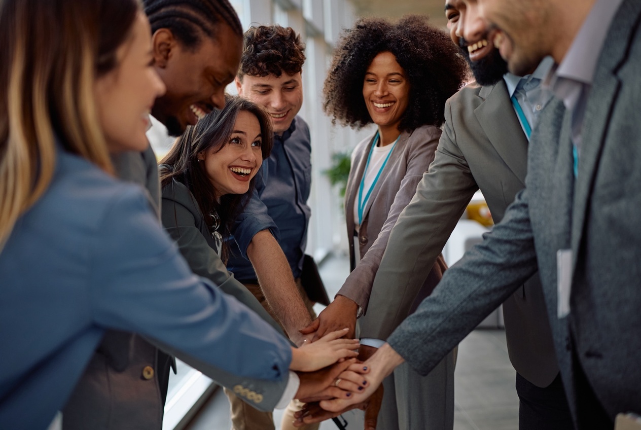 Diverse team of six adults in business attire stacking hands in the center to show teamwork and celebration in an office setting.
