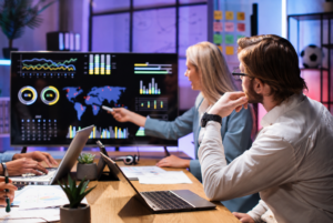 Team of analysts studying a data dashboard on a large monitor, with graphs, charts, and a world map in a modern office setting.
