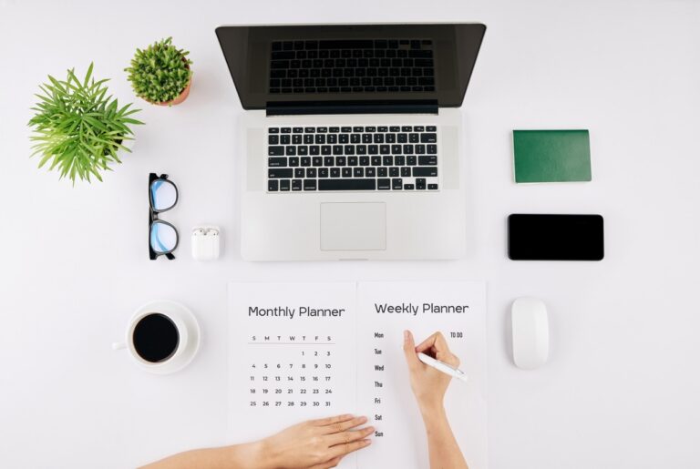 Top-down view of a minimalist workspace: laptop, planners, coffee, plants, glasses, phone, and accessories on a white desk.