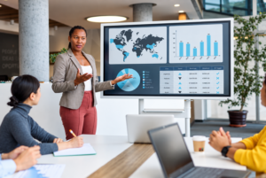 Presenter explains a data dashboard with a world map, bar and pie charts to a team in a modern meeting room.