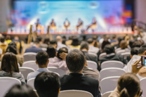 Audience seated in a conference hall watching a panel discussion on a brightly lit stage.