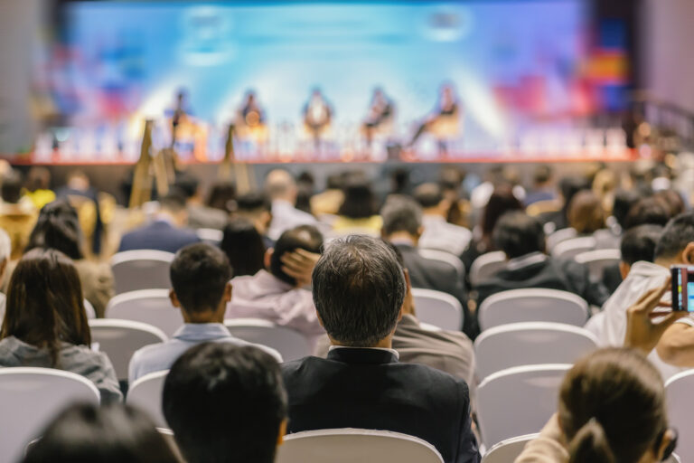 Audience seated in a conference hall watching a panel discussion on a brightly lit stage.